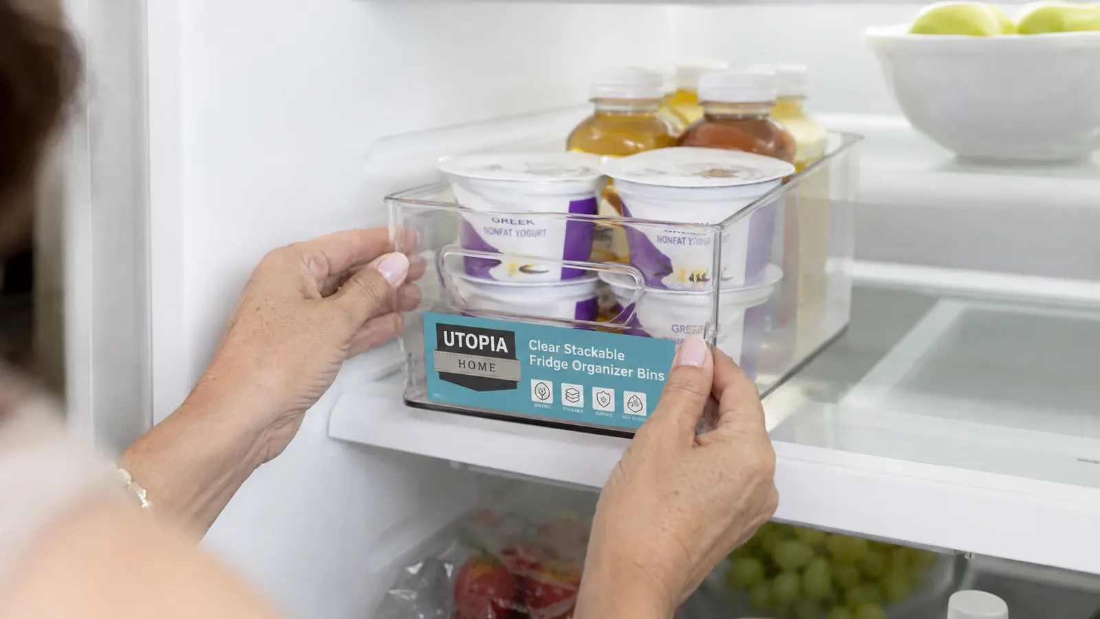 Hands sliding a clear fridge organizer bin onto a refrigerator shelf filled with yogurt cups and condiments