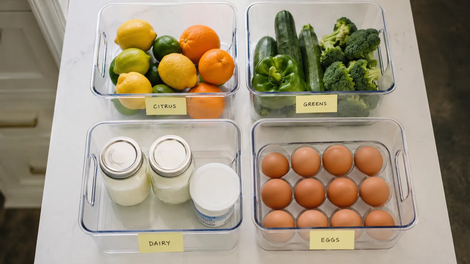 Clear fridge bins grouped by category, one labeled snacks, one labeled leftovers, one labeled produce, viewed from above